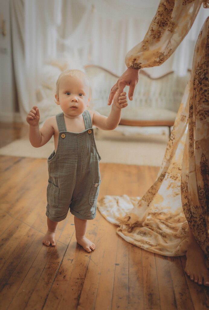 Blonde young boy standing up, wearing a textured blue overalls. He is holding his mothers hand.