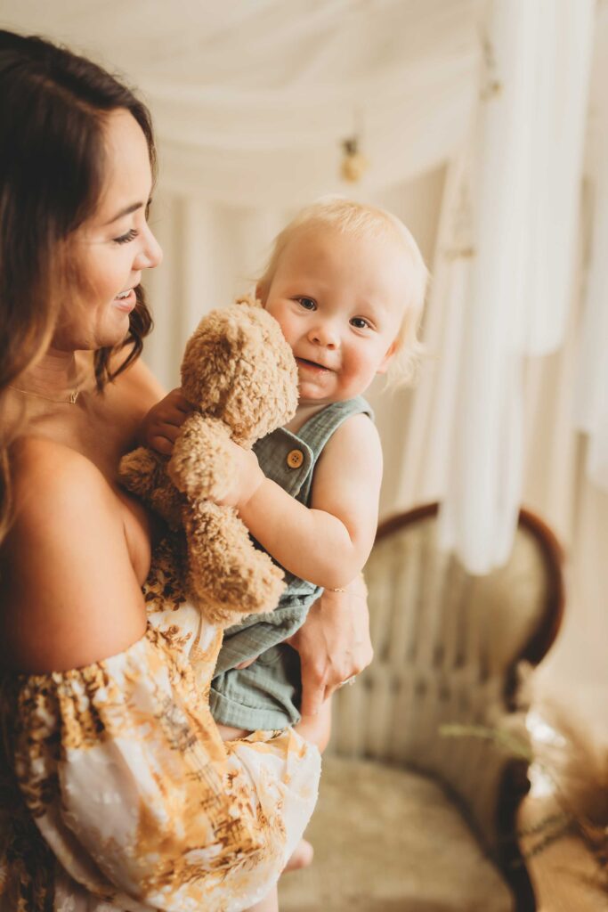 Baby boy holding a fuzzy teddy bear. He is smiling while his mother holds him and looks at him.