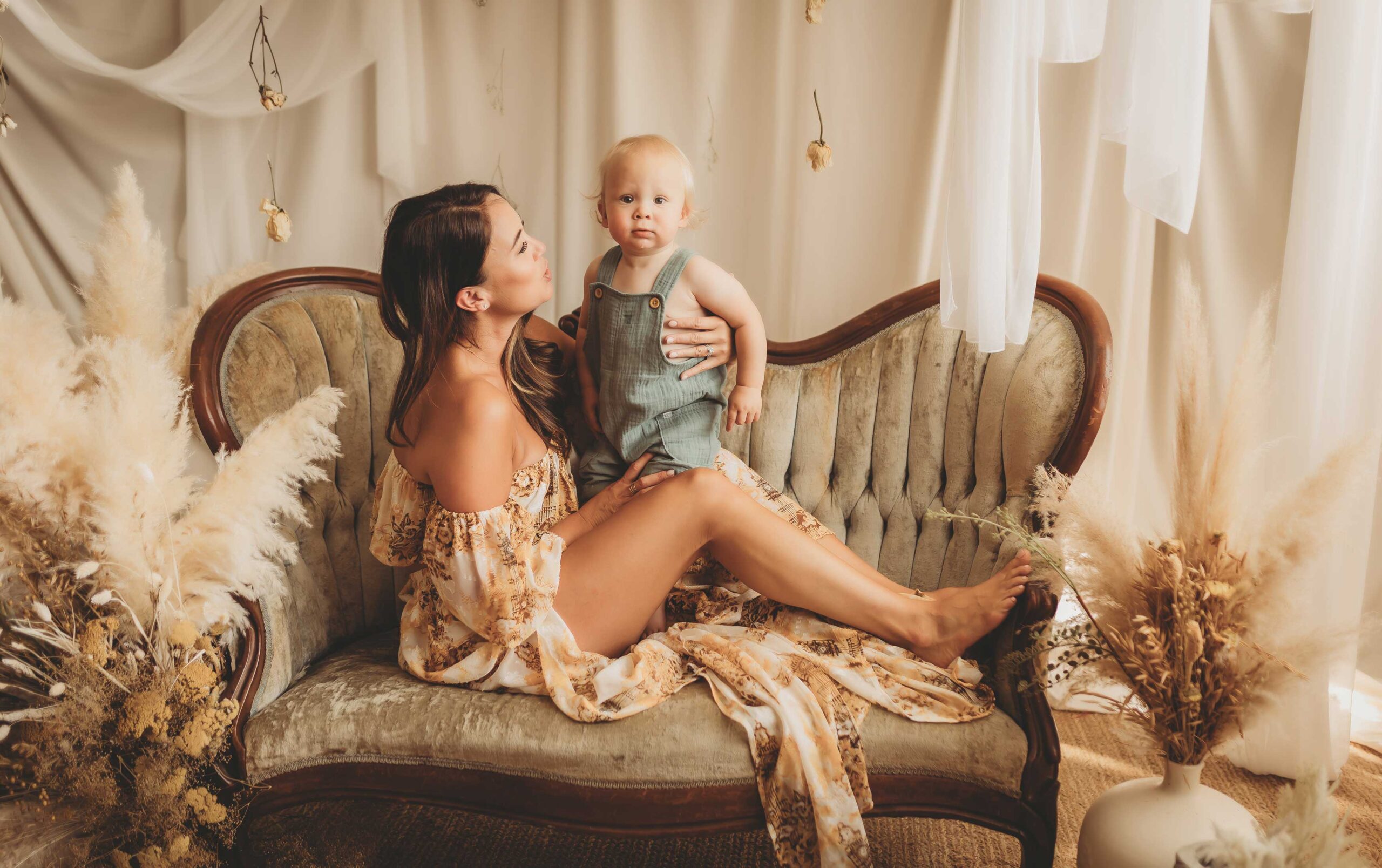 Mother sitting on a velvet sofa wearing a white and yellow dress while she holds her baby boy.