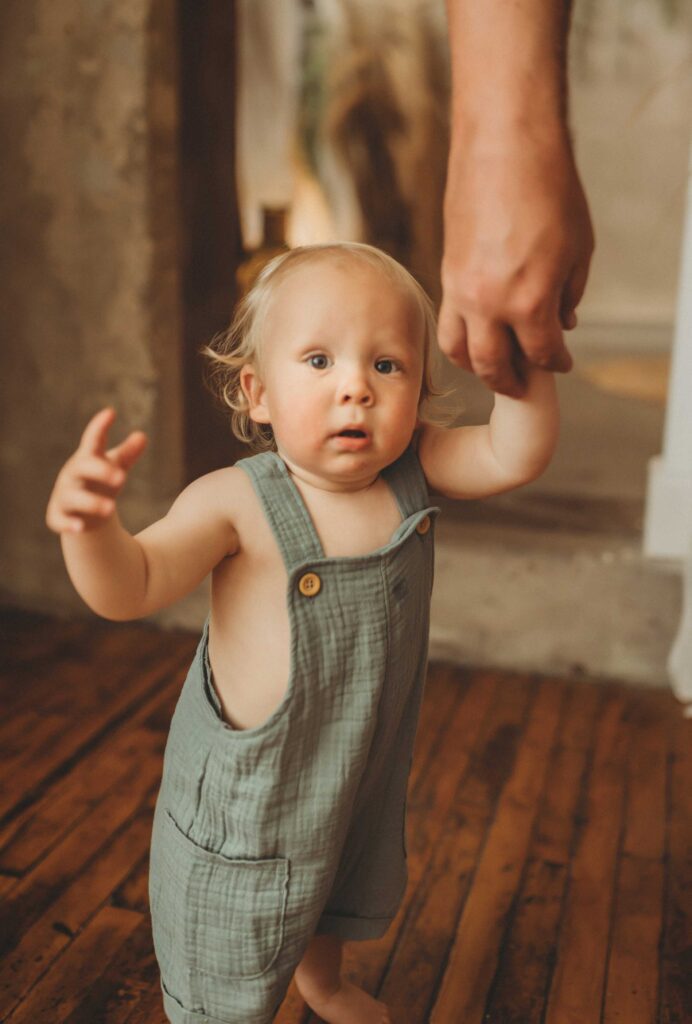 Blonde boy looking straight forward pointing with his right hand and holding his mother's hand with his left hand.