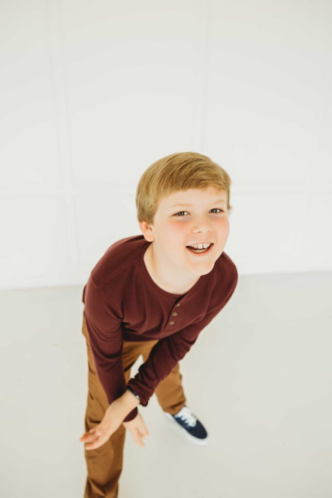 Young red haired boy smiling and moving his arms. He is wearing a burgundy henley t-shirt and a tan colored pant