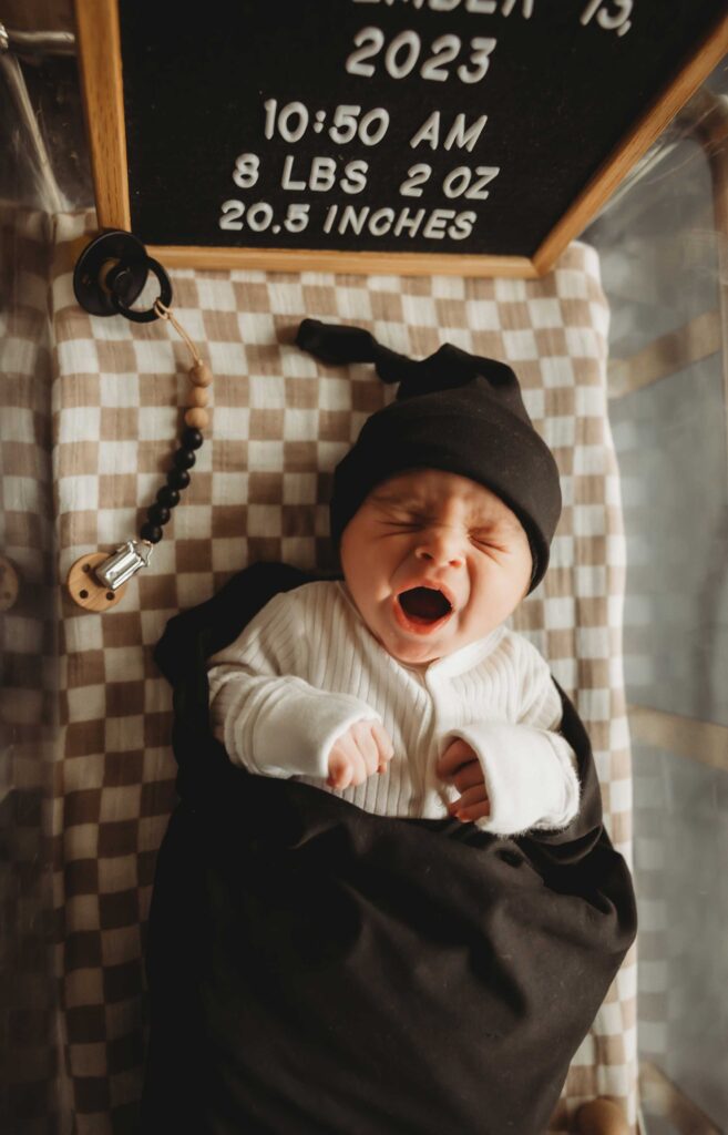newborn baby yawning and resting on a hospital crib. he is wearing a black knitted hat and a white onesie