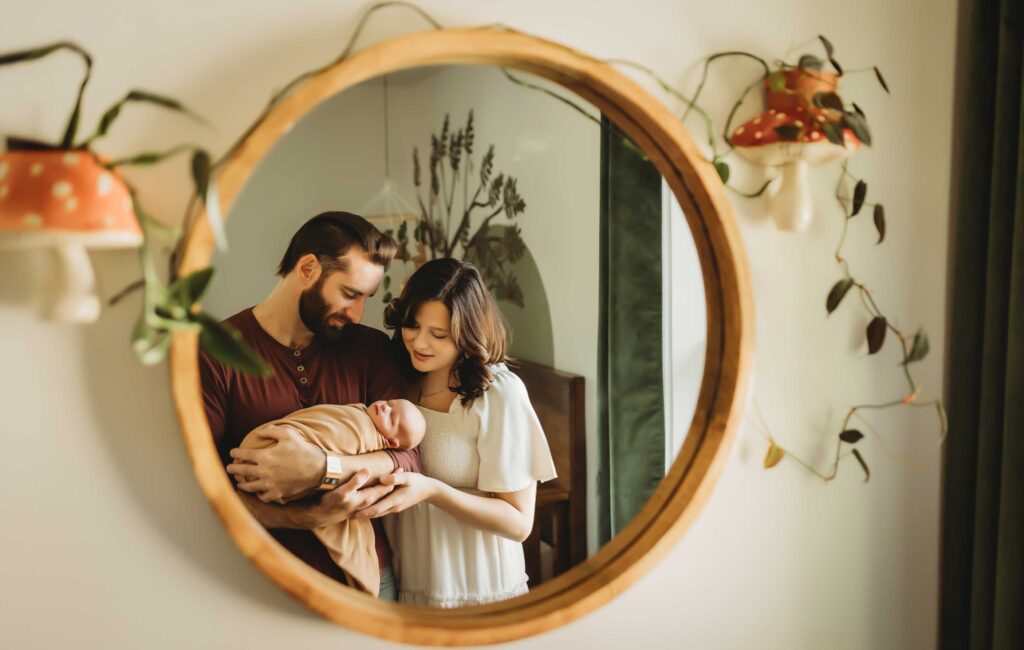 Reflection of a mother and father in a round mirror. The mirror has a wood frame and mushroom shade lamps on the sides