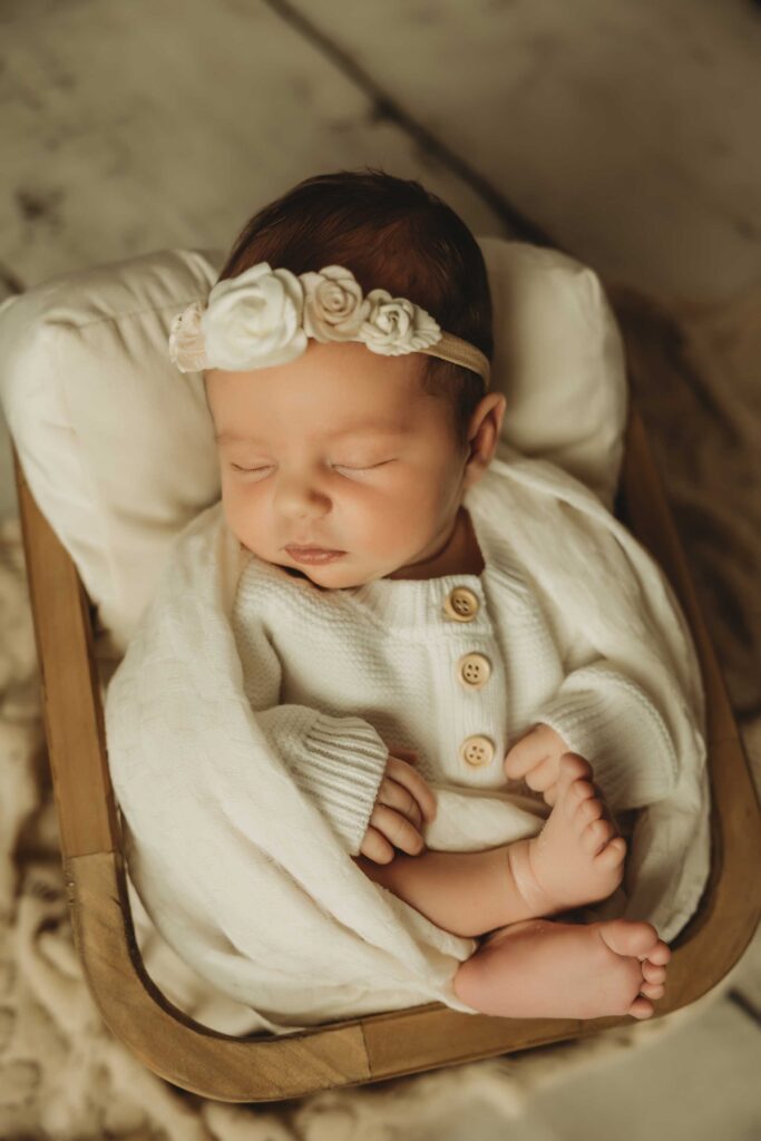 Baby sleeping in a toy wooden crib wearing a white onesie and a white headband with flowers