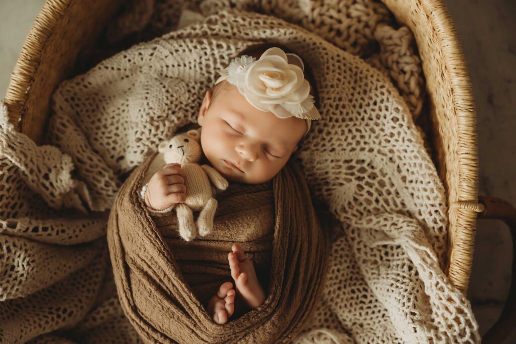 Baby girl sleeping in a basket, holding a small white teddy bear. She is wrapped in a brown wrap and wearing a white flower headband