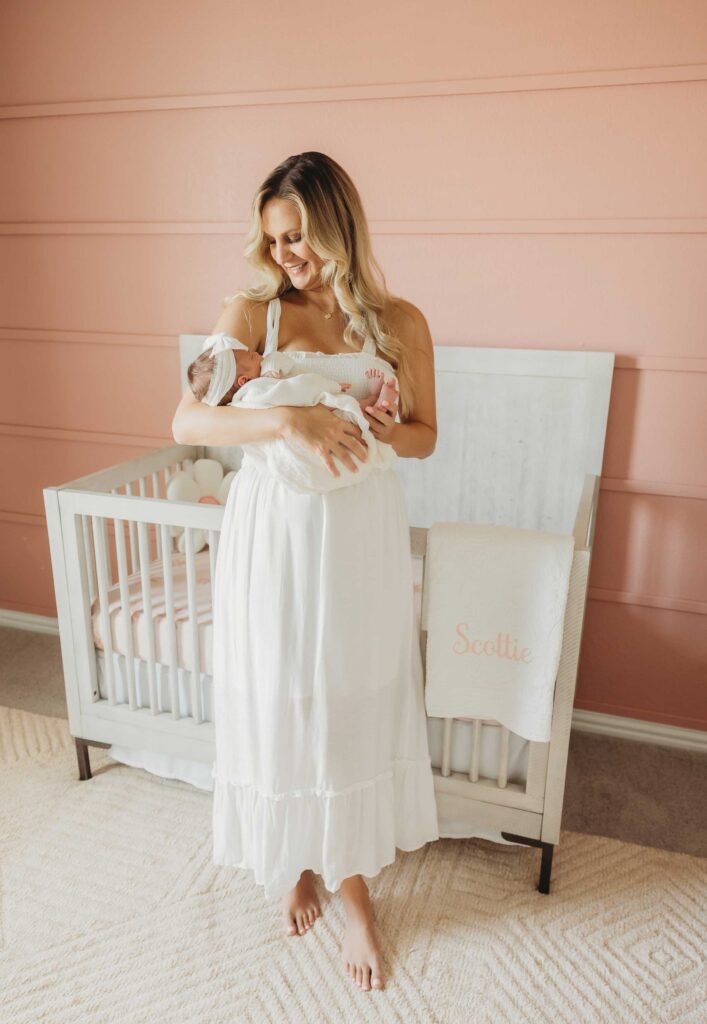 Blond woman standing in the middle of her daughter's nursery holding her baby. In the background you can see a white crib and a pink wall.