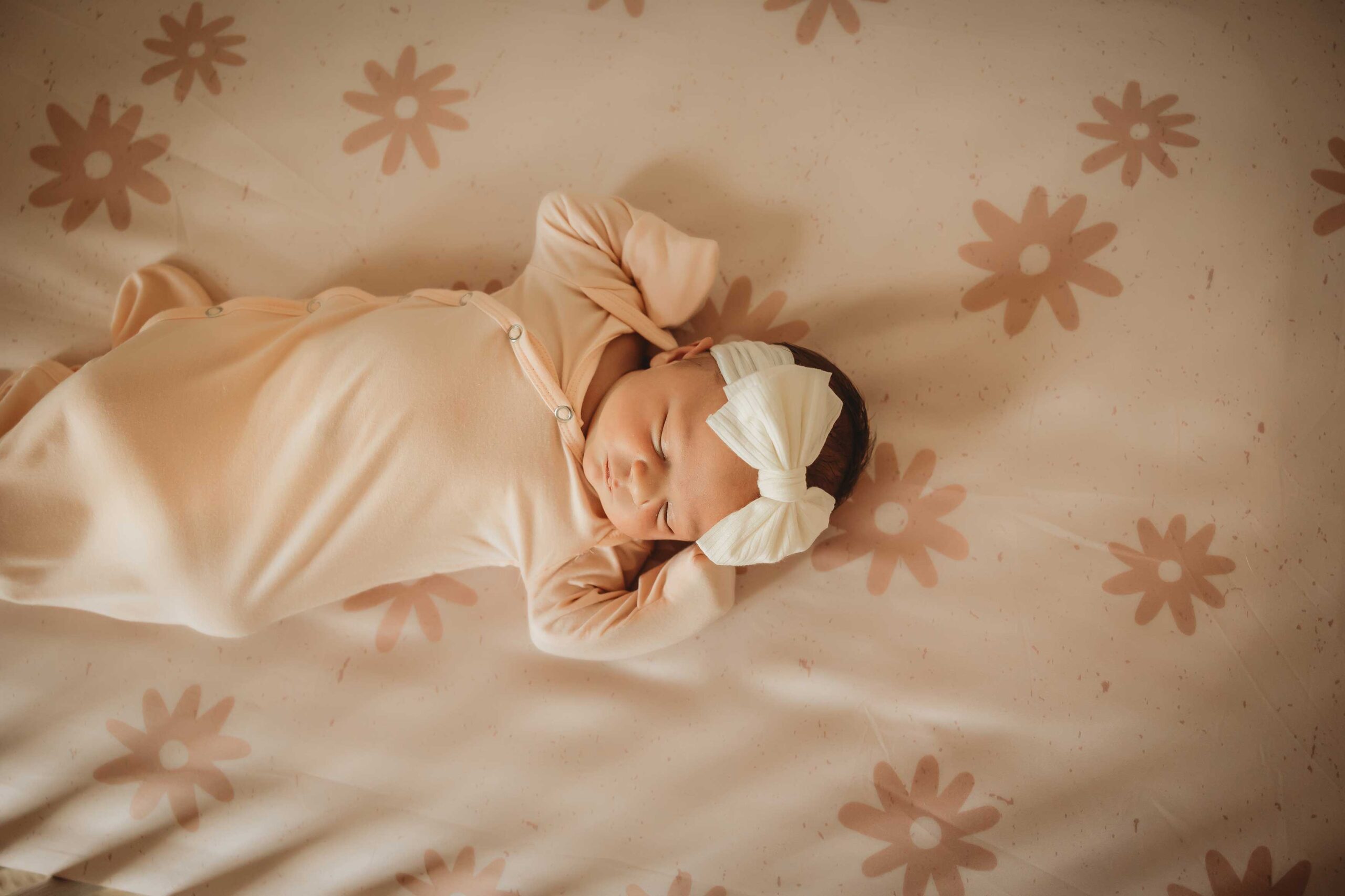 Sweet baby girl wearing a pink onesie and a white headband with a big bow. She is laying down on a pink flower sheet