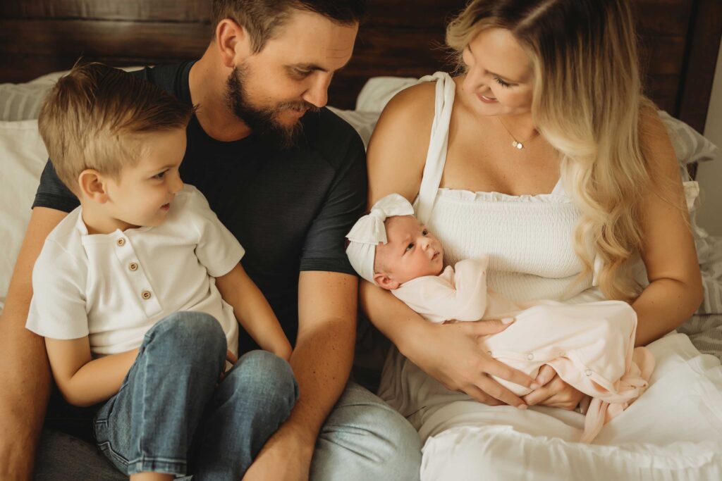 Couple sitting on a bed while holding her new baby girl and their son who is wearing a white shirt and jeans