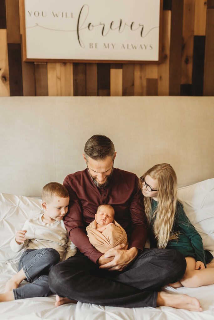 Father sitting on a bed with his three kids smiling and looking at the newborn baby