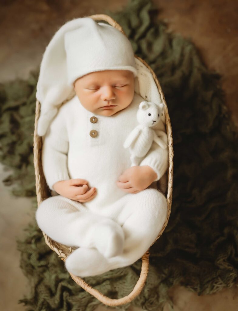 Baby sleping in a small basket wearing a white onesie and a white soft hat. He is holding a small white teddy bear.