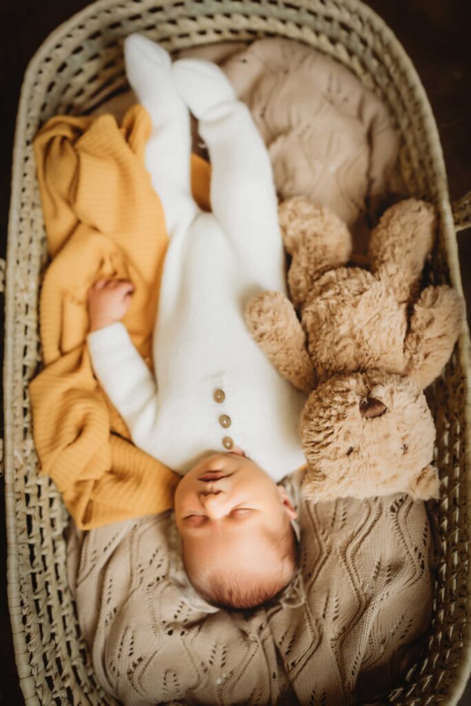 Baby sleeping in a basket wearing a white cozy onesie. Besides him is a fluffy brown teddy bear