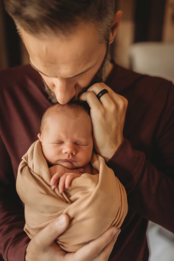 Man kissing baby on the top of his head. The baby is sleeping and is wrapped in a soft textured blanket.