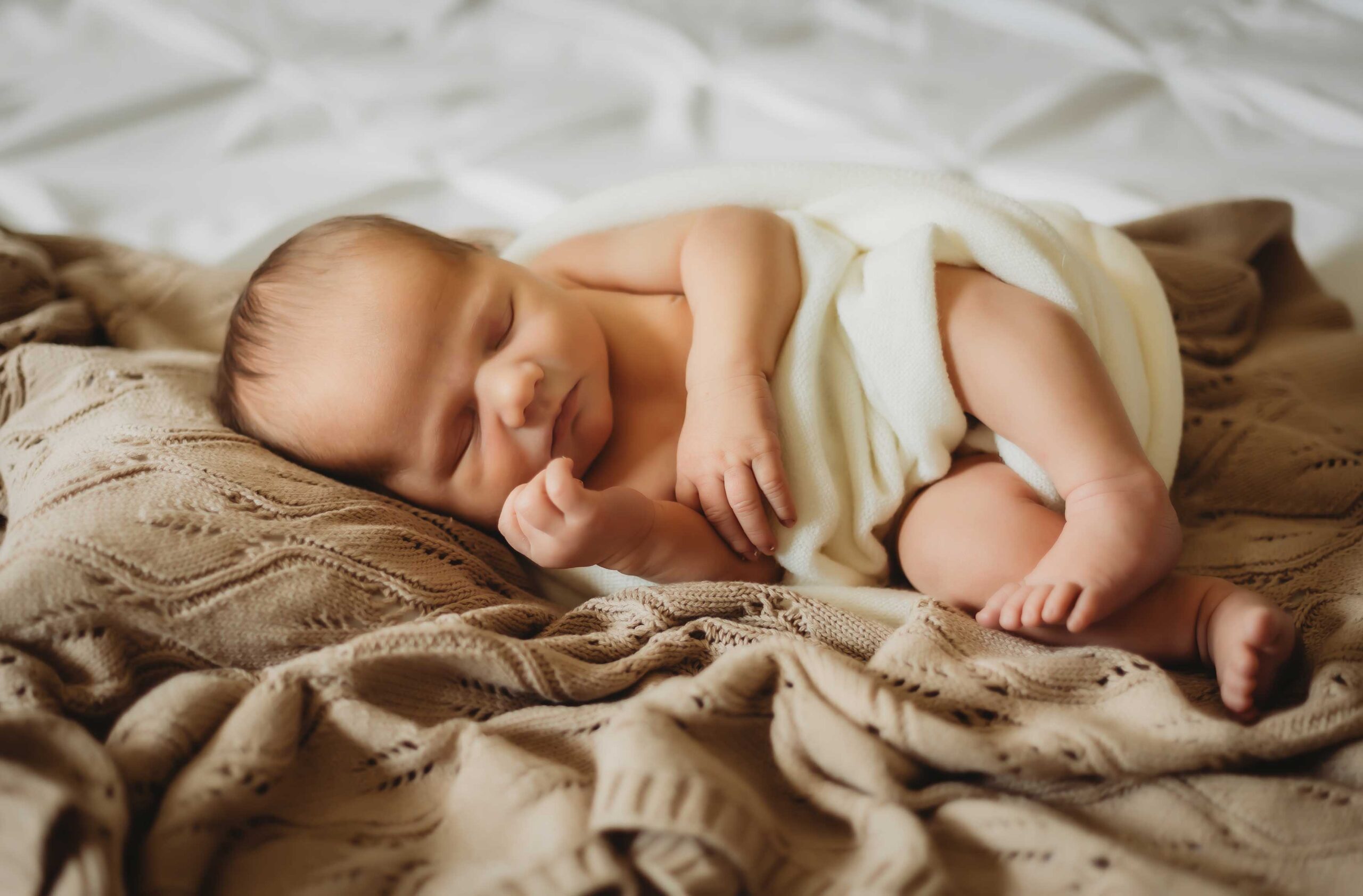 Newborn baby sleeping on a bed wearing nothing but a white soft blanket around his diaper. in home newborn session