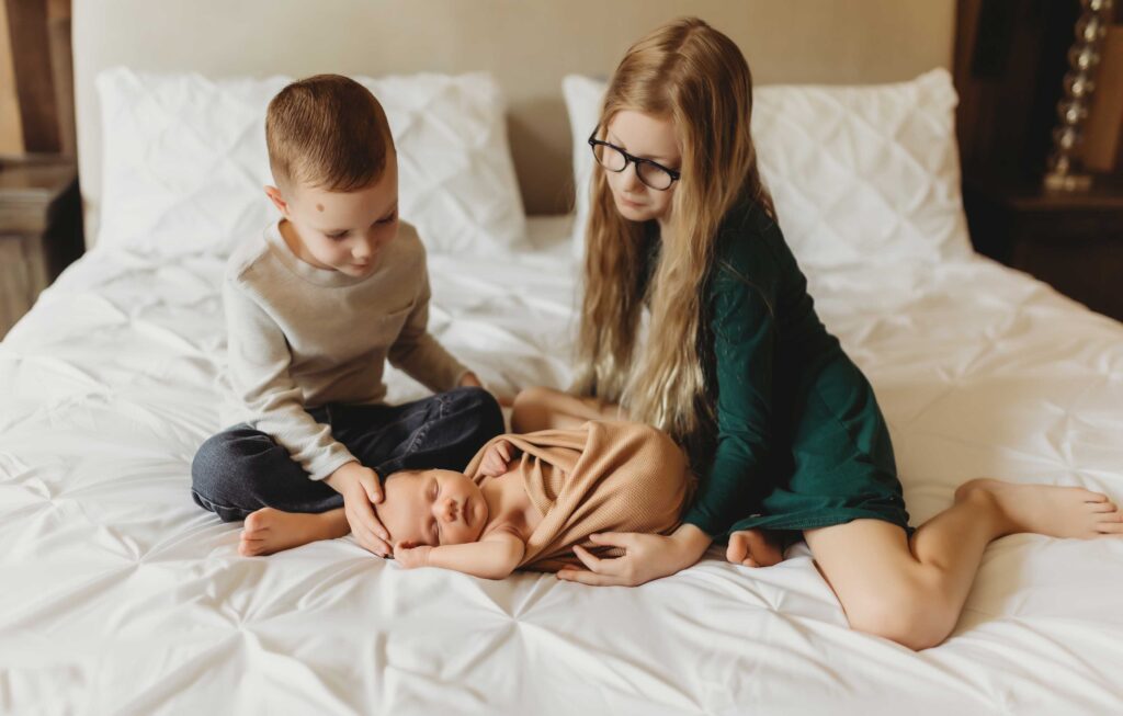 Girl wearing a green dress and glasses and boy wearing a T-shirt and jeans are sitting on a bed looking at their newborn baby sister